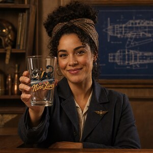 Op de afbeelding: Een helder glazen drinkglas met een afbeelding van een zwarte kat met een pilotenhelm en een bril, boven de tekst Nine Lives in een gestileerd geel lettertype. Het glas heeft een taps toelopende cilindrische vorm met een gladde afwerking.