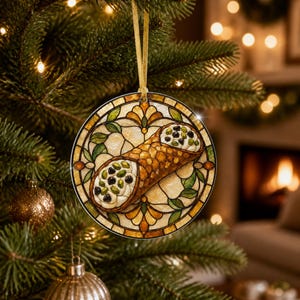 May include: A round, flat ornament featuring a stained-glass style illustration of a cannoli pastry with a golden-brown shell and white cream filling with dark chocolate chips, suspended by a thin gold ribbon against a green pine tree background.