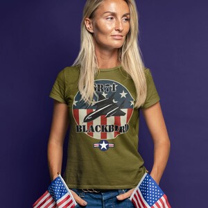 May include: Olive green t-shirt featuring a graphic of the SR-71 Blackbird aircraft with a red, white, and blue design. The text "SR-71 BLACKBIRD" is displayed on the shirt. The person is wearing blue jeans and holding two American flags.