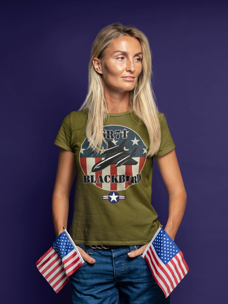 May include: Olive green t-shirt featuring a graphic of the SR-71 Blackbird aircraft with a red, white, and blue design. The text "SR-71 BLACKBIRD" is displayed on the shirt. The person is wearing blue jeans and holding two American flags.
