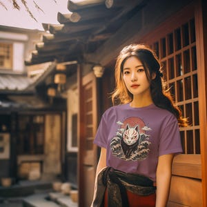 May include: A short-sleeved purple t-shirt featuring a graphic print of a white fox mask with red markings, positioned in front of a large red sun and stylised dark blue ocean waves. The design is centred on the chest of the garment.