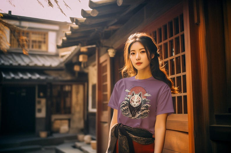 May include: A short-sleeved purple t-shirt featuring a graphic print of a white fox mask with red markings, positioned in front of a large red sun and stylised dark blue ocean waves. The design is centred on the chest of the garment.