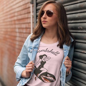 May include: A light pink t-shirt with the word "Bombshell" in script and a vintage-style illustration of a woman riding a shark. The person is wearing a light blue denim jacket and sunglasses.