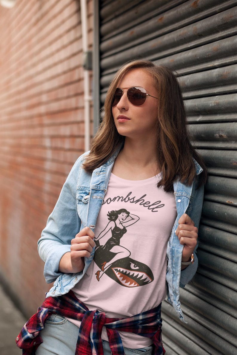 May include: A light pink t-shirt with the word "Bombshell" in script and a vintage-style illustration of a woman riding a shark. The person is wearing a light blue denim jacket and sunglasses.