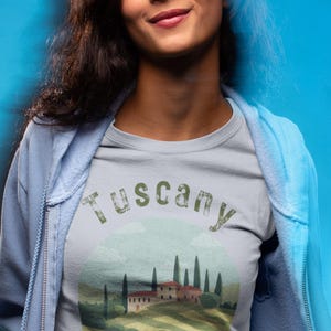 May include: A light gray t-shirt with a Tuscany landscape design. The shirt features a scenic illustration of a Tuscan villa, cypress trees, and rolling hills. The text "Tuscany" and "Italia Bella" are printed on the shirt.