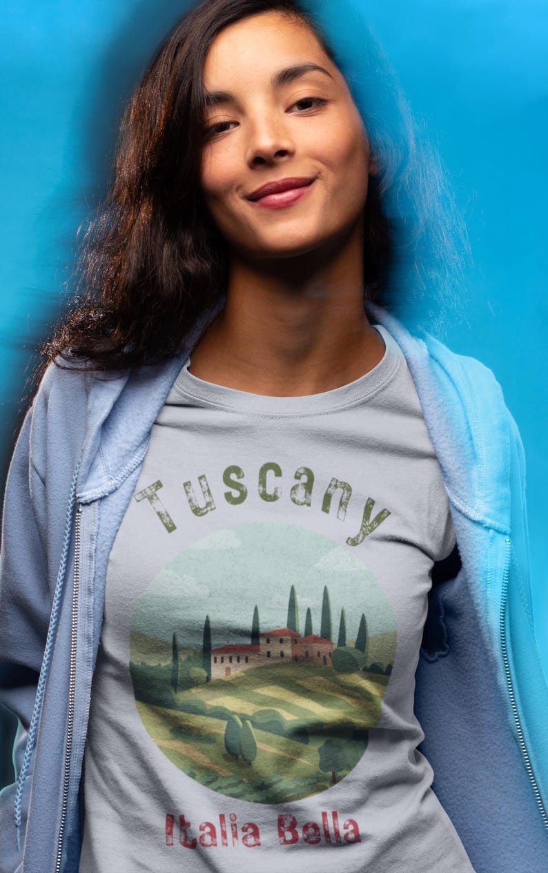 May include: A light gray t-shirt with a Tuscany landscape design. The shirt features a scenic illustration of a Tuscan villa, cypress trees, and rolling hills. The text "Tuscany" and "Italia Bella" are printed on the shirt.