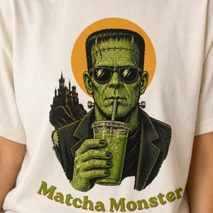 Może przedstawiać: Kremowy t-shirt z grafiką przedstawiającą zielonego potwora w okularach przeciwsłonecznych i ciemnej marynarce, pijącego zielony nap&oacute;j z przezroczystego kubka ze słomką. Za postacią widoczny jest zarys zamku oraz ż&oacute;łte koło z napisem Matcha Monster.