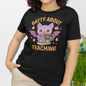 K&ouml;nnte beinhalten: Schwarzes T-Shirt mit einem lila Fledermaus-Motiv, das eine Brille tr&auml;gt und eine Tafel mit "A+" h&auml;lt. Der Text "BATTY ABOUT TEACHING" ist &uuml;ber der Fledermaus. Das T-Shirt hat einen Rundhalsausschnitt und kurze &Auml;rmel.