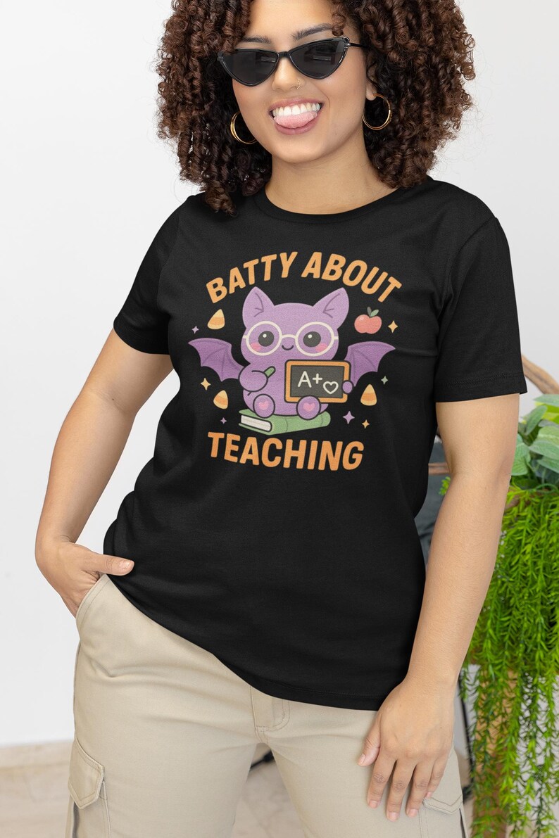 K&ouml;nnte beinhalten: Schwarzes T-Shirt mit einem lila Fledermaus-Motiv, das eine Brille tr&auml;gt und eine Tafel mit "A+" h&auml;lt. Der Text "BATTY ABOUT TEACHING" ist &uuml;ber der Fledermaus. Das T-Shirt hat einen Rundhalsausschnitt und kurze &Auml;rmel.