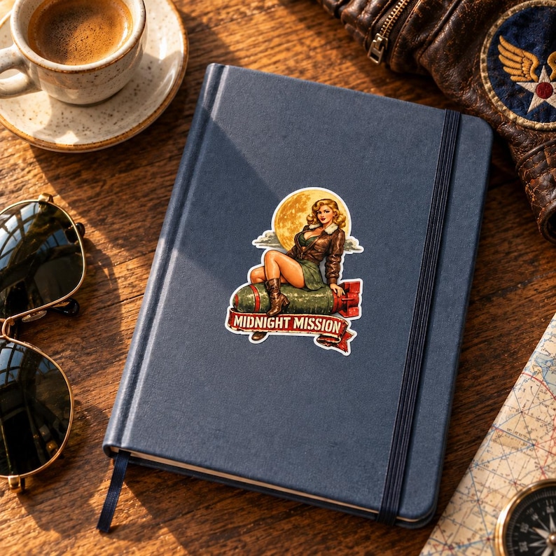 K&ouml;nnte beinhalten: Ein dunkelblaues Notizbuch mit einem Aufkleber einer blonden Pin-up-Figur in brauner Fliegerjacke, die auf einer gr&uuml;nen Bombe vor einem gelben Mond sitzt, mit einem roten Banner unten mit dem Text Midnight Mission.