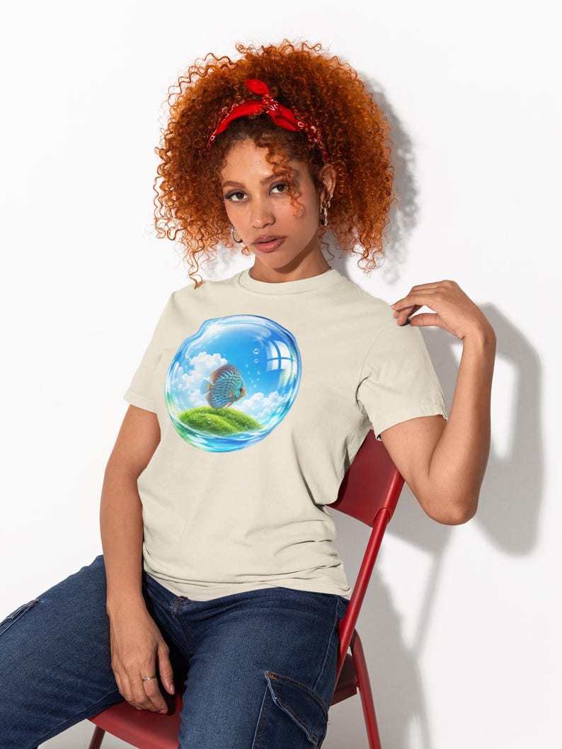 May include: A beige short-sleeved t-shirt featuring a graphic print of a blue fish swimming inside a transparent bubble that contains a small grassy island under a bright blue sky with white clouds and rising air bubbles.