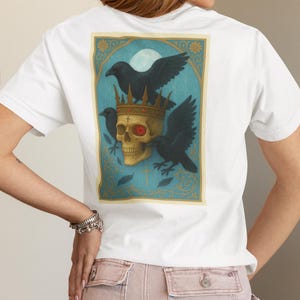 May include: White t-shirt with a back graphic of a crowned skull with a red eye, two black ravens, and a cross, set against a teal and gold background. The shirt has a crew neck design, a popular style in the UK.
