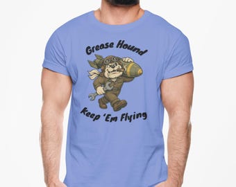 May include: Light blue t-shirt with the text "Grease Hound Keep 'Em Flying". The shirt features a cartoon dog in a pilot's outfit holding a wrench and a bomb. The dog is brown and the bomb is green and yellow.