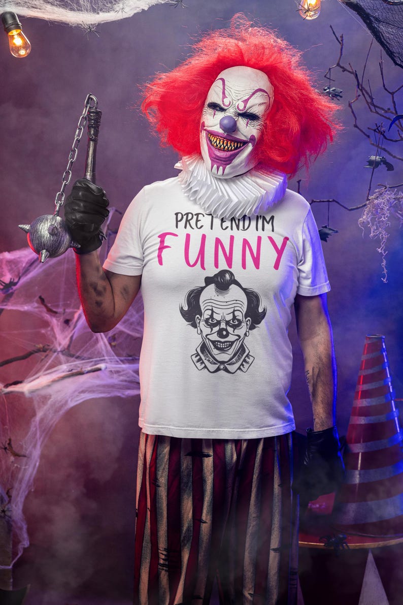 May include: A person in a clown mask and red wig, wearing a white t-shirt with the words "PRETEND IM FUNNY" in pink. The shirt also features a clown graphic. The person is holding a spiked mace and wearing black gloves. The background includes cobwebs and a traffic cone.