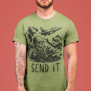 Puede incluir: Camiseta verde oliva con una ilustraci&oacute;n en blanco y negro de un soldado esqueleto con una radio, rodeado de otros soldados y aviones de combate. El texto "SEND IT" est&aacute; impreso debajo del gr&aacute;fico.