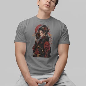 May include: A grey short-sleeved t-shirt featuring a graphic print of a person with dark hair wearing a black and red patterned garment, holding a fox mask, with red circular accents and floral details in the background of the illustration.