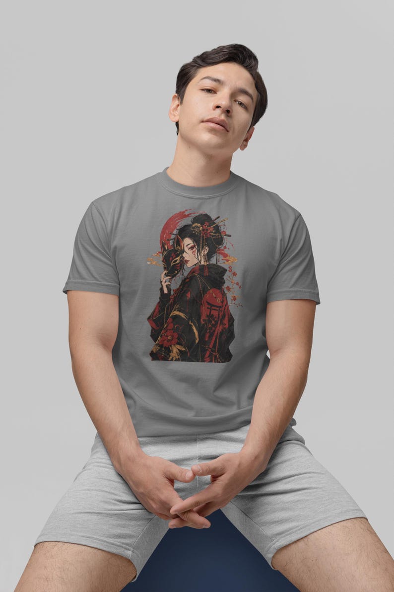 May include: A grey short-sleeved t-shirt featuring a graphic print of a person with dark hair wearing a black and red patterned garment, holding a fox mask, with red circular accents and floral details in the background of the illustration.