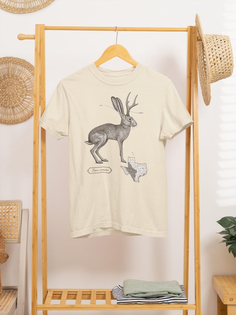 May include: A cream-colored short-sleeve t-shirt featuring a black and white ink-style illustration of a jackalope. Below the animal is the text Lepus cornutus and a map of Texas with a grid pattern. The shirt has a crew neckline and hem.