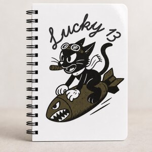 May include: A white spiral-bound notebook featuring a black and white graphic of a cartoon cat wearing goggles and a scarf while riding a bomb. The cat holds a cigar in its mouth. The text Lucky 13 is printed in a cursive black font above the cat.