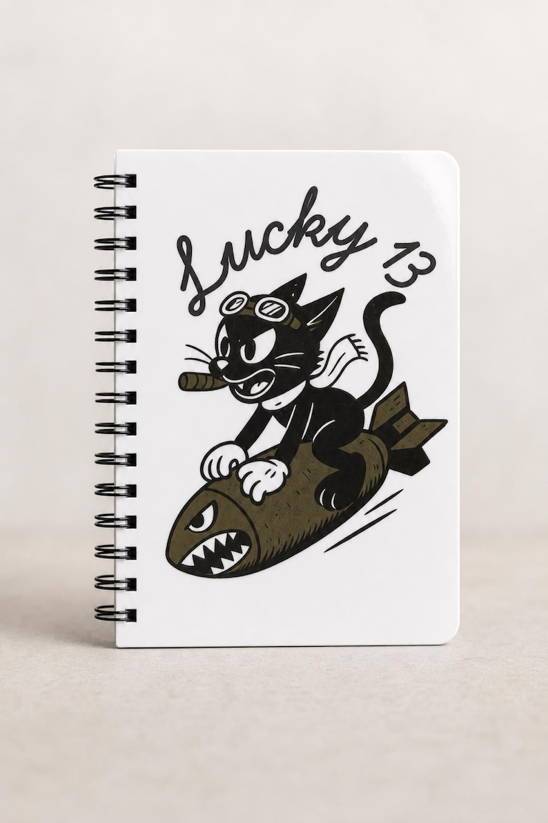 May include: A white spiral-bound notebook featuring a black and white graphic of a cartoon cat wearing goggles and a scarf while riding a bomb. The cat holds a cigar in its mouth. The text Lucky 13 is printed in a cursive black font above the cat.