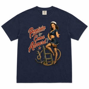 Op de afbeelding: Een marineblauw t-shirt met korte mouwen en een afbeelding van een pin-up zeeman die op een houten ladder staat naast een anker en touw. De tekst "Permission to Come Aboard?" staat in rode en witte letters aan de linkerkant gedrukt.
