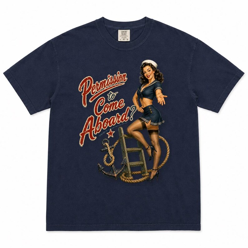 Op de afbeelding: Een marineblauw t-shirt met korte mouwen en een afbeelding van een pin-up zeeman die op een houten ladder staat naast een anker en touw. De tekst "Permission to Come Aboard?" staat in rode en witte letters aan de linkerkant gedrukt.