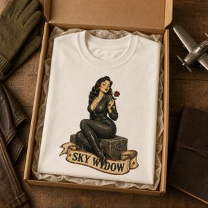 May include: A white t-shirt featuring a graphic of a woman with dark hair wearing a black jumpsuit, sitting on a wooden crate while holding a red rose. Below the crate is a banner with the text SKY WIDOW in bold, dark, capital letters.