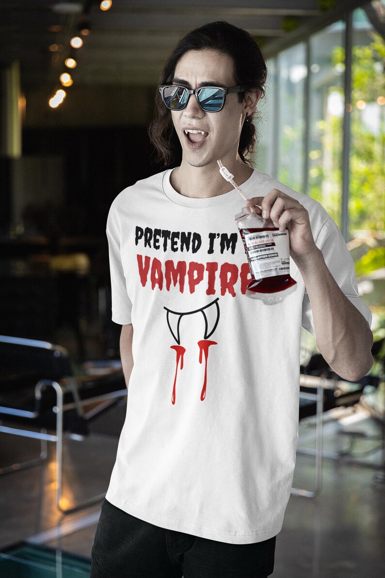 May include: White t-shirt with the text "PRETEND I'M VAMPIRE" in black and red. Below the text is a graphic of vampire fangs with red drips. The person is holding a drink in a blood bag.
