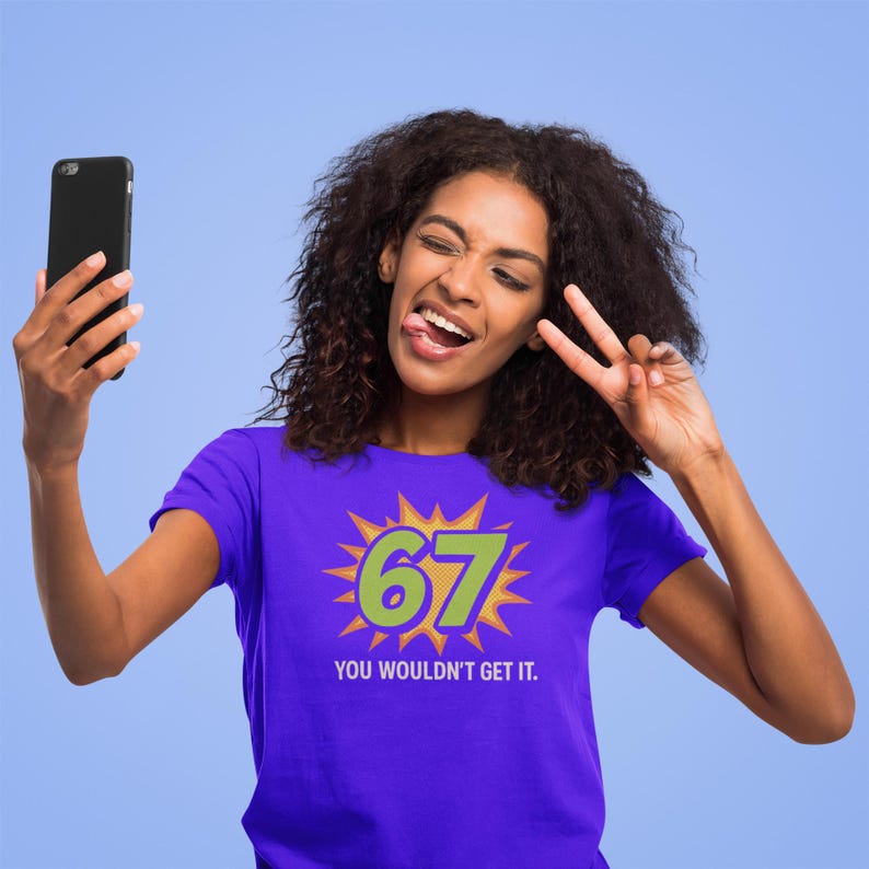 May include: A purple t-shirt with the number 67 in green and yellow, with the text "YOU WOULDN'T GET IT." A person is holding a black smartphone and making a peace sign.