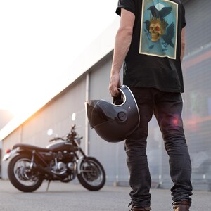 May include: Black t-shirt with a skull and raven graphic. A person holds a black motorcycle helmet. A black motorcycle is in the background. The person wears black jeans and brown boots.