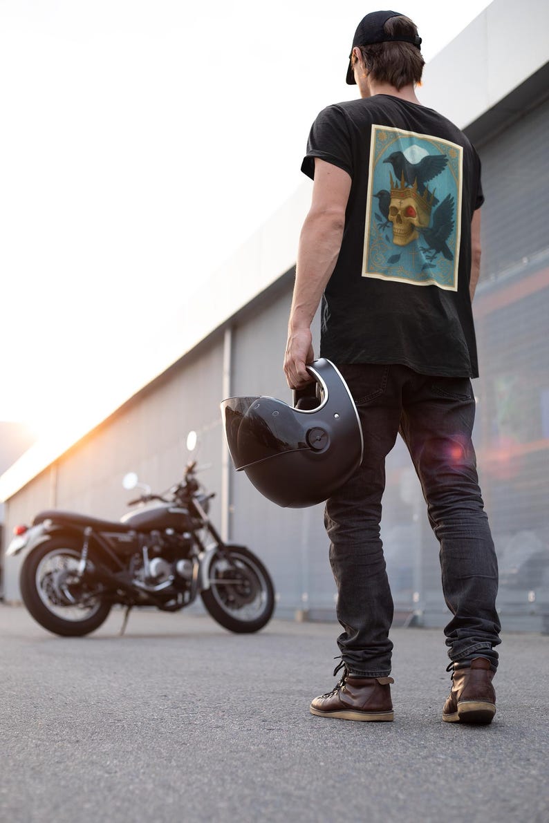 May include: Black t-shirt with a skull and raven graphic. A person holds a black motorcycle helmet. A black motorcycle is in the background. The person wears black jeans and brown boots.