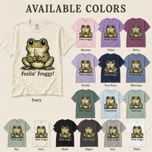 Może przedstawiać: Kolekcja koszulek z kr&oacute;tkim rękawem w r&oacute;żnych kolorach, każda z grafiką żaby trzymającej n&oacute;ż i napisem 'Feelin' Froggy!' poniżej. Dostępne kolory to kość słoniowa, blossom, fioletowy, berry, orchid, true navy, blue jean, blue spruce, chambray, seafoam, bay, czarny, pepper, szary i biały.