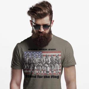 May include: A grey t-shirt featuring a graphic of seven skeleton soldiers in various historical military uniforms standing in front of a faded American flag. The text "DEAD MEN STILL" appears above the soldiers and "Stand for the Flag" below.