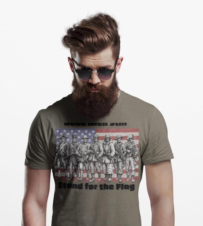 May include: A grey t-shirt featuring a graphic of seven skeleton soldiers in various historical military uniforms standing in front of a faded American flag. The text "DEAD MEN STILL" appears above the soldiers and "Stand for the Flag" below.