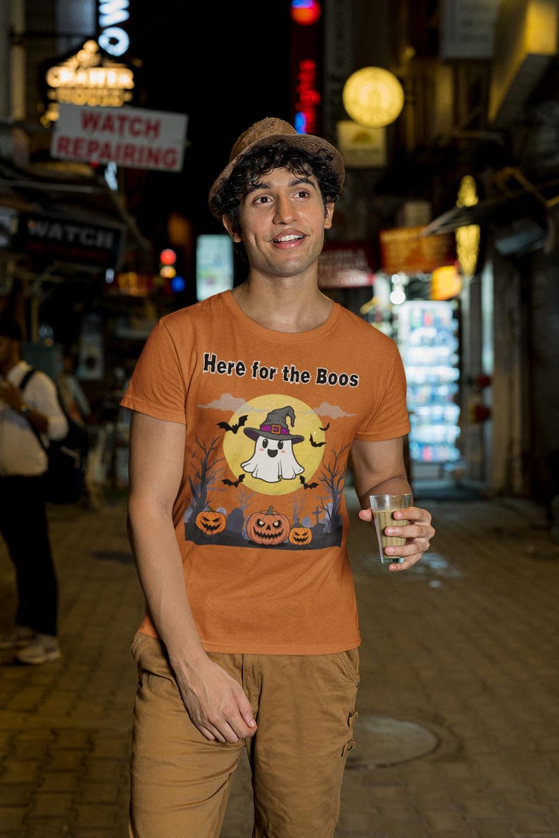 May include: Orange t-shirt with the text "Here for the Boos" and a Halloween graphic featuring a ghost, bats, and pumpkins. The person is wearing a brown hat and khaki pants.