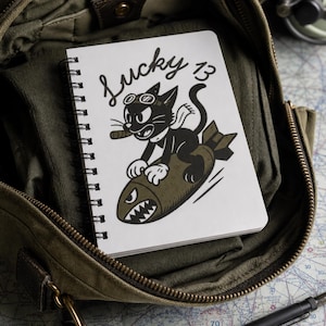 May include: A white spiral-bound notebook featuring a black and white illustration of a cartoon cat wearing goggles and a scarf, riding a brown bomb with a shark-mouth design. The text Lucky 13 is printed in black script above the cat.