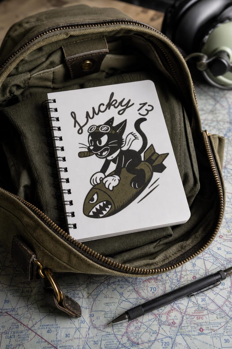May include: A white spiral-bound notebook featuring a black and white illustration of a cartoon cat wearing goggles and a scarf, riding a brown bomb with a shark-mouth design. The text Lucky 13 is printed in black script above the cat.