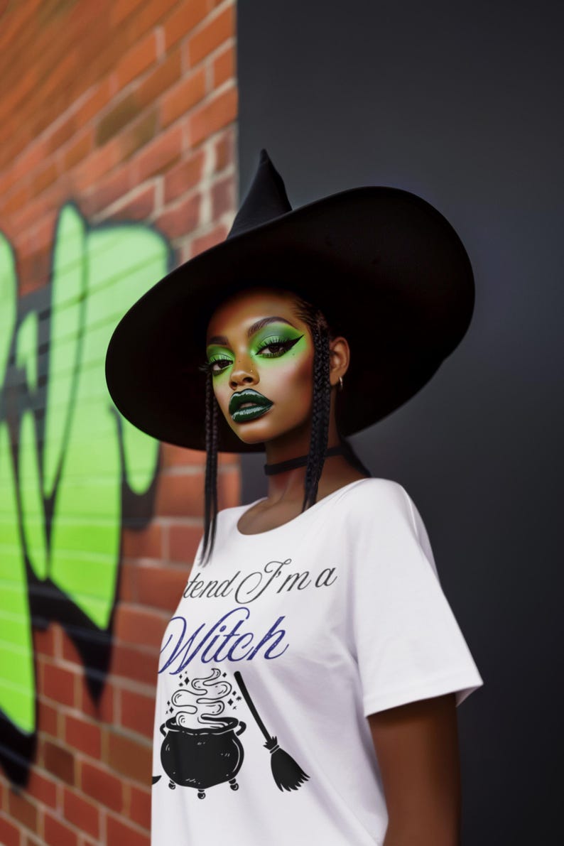 May include: A person wearing a black witch hat and a white t-shirt with the text "I pretend I'm a Witch". The person has green eye makeup and dark green lipstick. The background includes a brick wall and green graffiti.