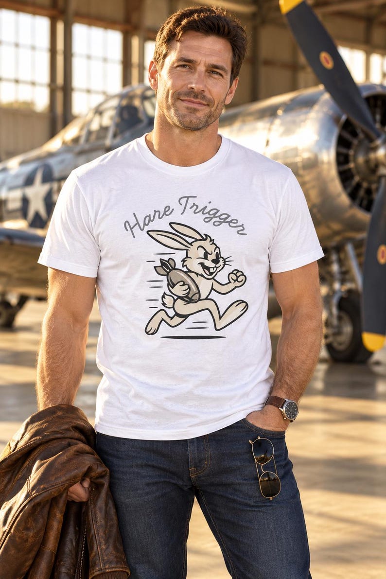 Puede incluir: Una camiseta blanca de manga corta con un gr&aacute;fico de un conejo de dibujos animados corriendo mientras sostiene una bomba. Sobre el conejo, el texto "Hare Trigger" est&aacute; impreso en una fuente cursiva oscura. El dise&ntilde;o usa tonos beige, marr&oacute;n y negro.