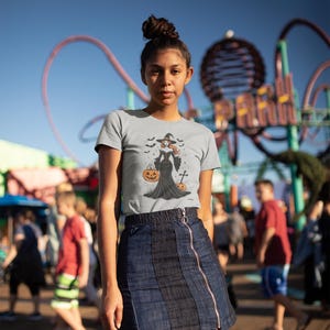 May include: A light gray t-shirt with a Halloween witch graphic, paired with a dark blue denim skirt with a front zipper. The witch graphic includes a witch, bats, and pumpkins. The background shows a blurred amusement park scene.