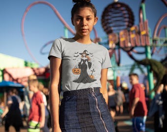 May include: A light grey t-shirt featuring a Halloween witch design, worn with a dark blue denim skirt with a front zip. The witch graphic includes a witch, bats, and pumpkins. The background shows a blurred amusement park scene.