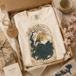 May include: A cream-coloured t-shirt featuring a graphic of a nine-tailed fox sitting under a cherry blossom tree with a full moon in the background, all enclosed in an ornate archway frame. The shirt has a Comfort Colors brand label at the neck.