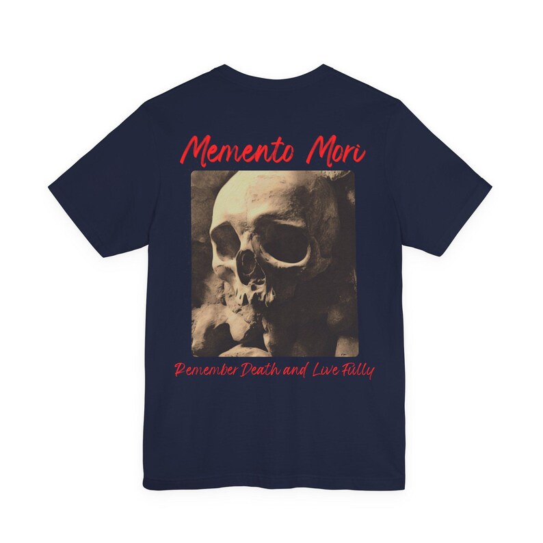 May include: Navy blue t-shirt with a skull graphic. The skull is in a sepia tone, with visible details. Above the skull is the text 'Memento Mori' in red, and below it, the text 'Remember Death and Live Fully' in red. The t-shirt is a basic crew neck.