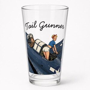 May include: A clear glass pint glass featuring a graphic illustration of a blue military aircraft with a pilot in the cockpit and a pin-up style figure operating a machine gun. The text Tail Gunner is printed in black script above the aircraft.
