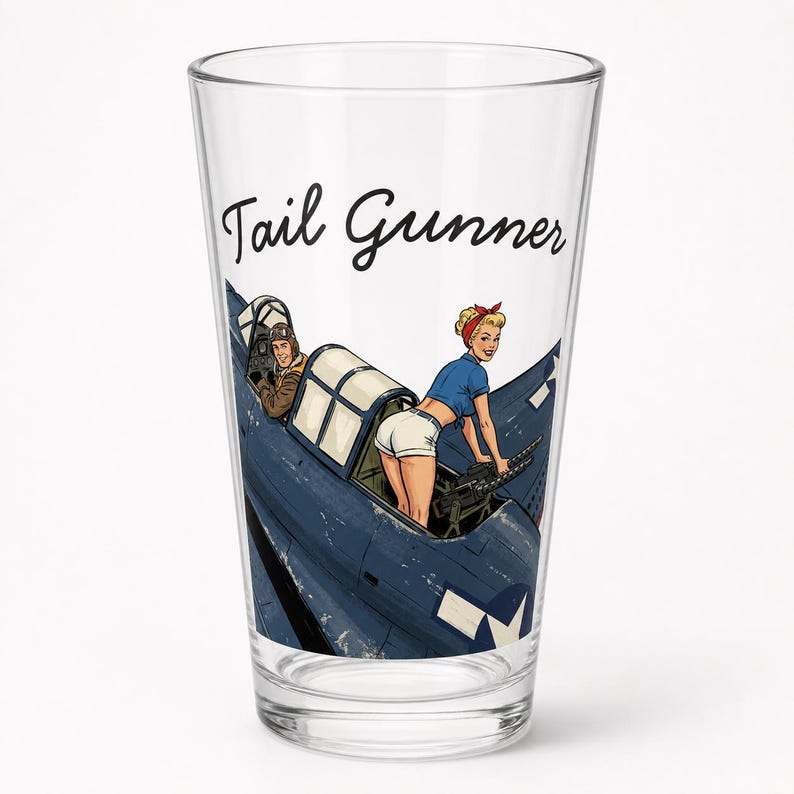 May include: A clear glass pint glass featuring a graphic illustration of a blue military aircraft with a pilot in the cockpit and a pin-up style figure operating a machine gun. The text Tail Gunner is printed in black script above the aircraft.