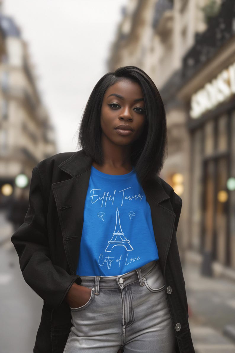 May include: A woman wearing a blue t-shirt with a white Eiffel Tower graphic and the words 'Eiffel Tower' and 'City of Love.' She is also wearing a black blazer and gray jeans. The background is a blurred street scene with buildings.