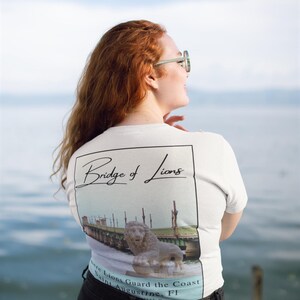 Peut inclure: T-shirt blanc avec un motif du pont des Lions &agrave; St. Augustine, Floride. Le design comprend une image d&eacute;taill&eacute;e du pont et d'une statue de lion, avec le texte "Bridge of Lions" et "Where Lions Guard the Coast".