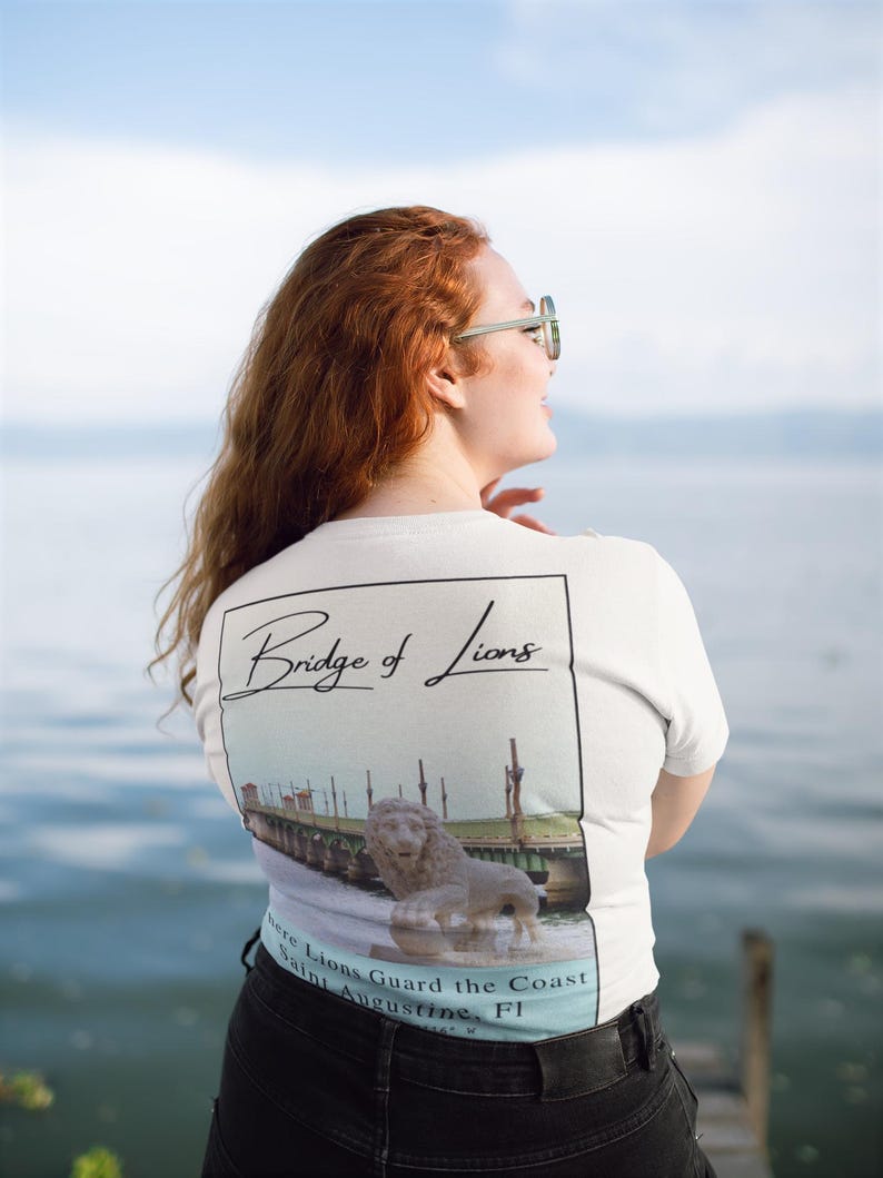 Peut inclure: T-shirt blanc avec un motif du pont des Lions &agrave; St. Augustine, Floride. Le design comprend une image d&eacute;taill&eacute;e du pont et d'une statue de lion, avec le texte "Bridge of Lions" et "Where Lions Guard the Coast".