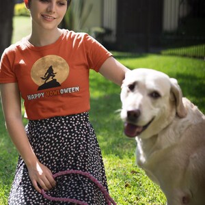 May include: An orange t-shirt with the text "HAPPY HOWLOWEEN" and a werewolf graphic against a full moon. The person wears a black and white polka dot skirt and holds a pink leash. A yellow Labrador Retriever sits on the grass.