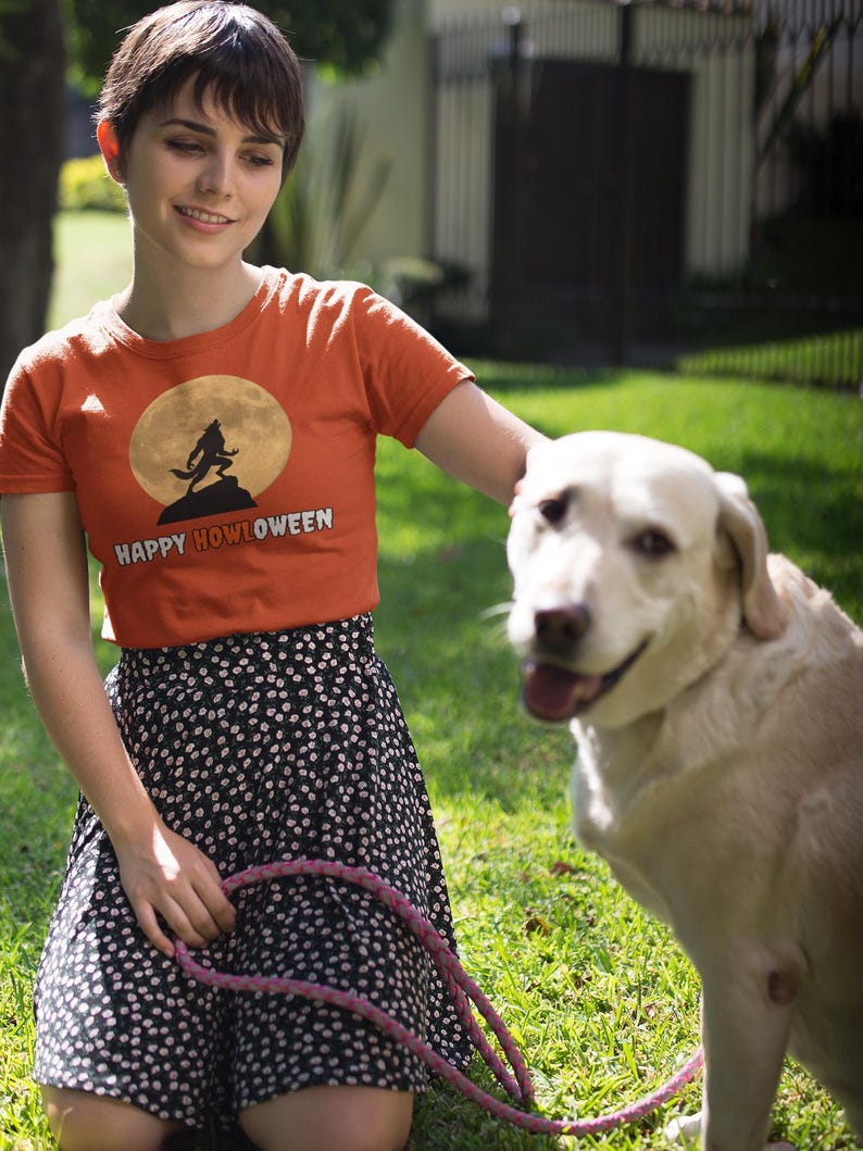 May include: An orange t-shirt with the text "HAPPY HOWLOWEEN" and a werewolf graphic against a full moon. The person wears a black and white polka dot skirt and holds a pink leash. A yellow Labrador Retriever sits on the grass.